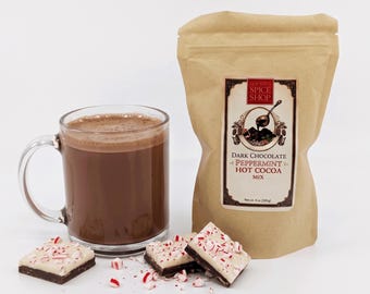 Peut inclure: Une tasse en verre transparent remplie de chocolat chaud brun foncé à côté d'un sachet en papier beige étiqueté Old Town Spice Shop Dark Chocolate Peppermint Hot Cocoa Mix, avec trois carrés de chocolat noir et blanc aux éclats de menthe.