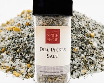 Op de afbeelding: Een doorzichtige glazen kruidenpot met een zwarte deksel gevuld met dille-augurkzout. Het etiket bevat de tekst Old Town Spice Shop, Dill Pickle Salt en het websiteadres. De pot staat voor een hoopje van het grove zoutmengsel.