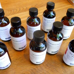 May include: A collection of ten small glass bottles with black screw-on caps. Each bottle contains a different type of extract, including anise, almond, orange, spearmint, wintergreen, and vanilla. The bottles are arranged in a semi-circle on a wooden surface.