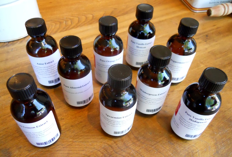 May include: A collection of ten small glass bottles with black screw-on caps. Each bottle contains a different type of extract, including anise, almond, orange, spearmint, wintergreen, and vanilla. The bottles are arranged in a semi-circle on a wooden surface.