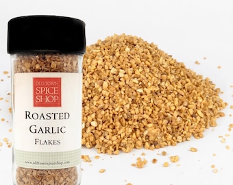 May include: A clear plastic spice jar with a black lid containing roasted garlic flakes. The label features the text Old Town Spice Shop Roasted Garlic Flakes and the website address. A pile of the same golden-brown garlic flakes sits beside.