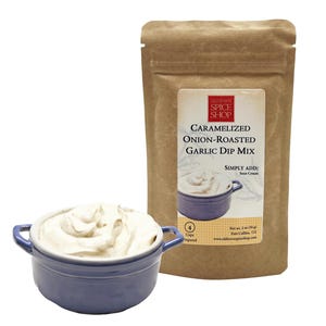 May include: A brown paper pouch with a label that reads "Caramelized Onion-Roasted Garlic Dip Mix" from Oldtown Spice Shop. The pouch is next to a blue ceramic bowl filled with a creamy white dip.