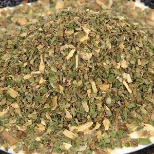May include: A mound of dried herb seasoning consisting of small, fragmented green leaves and light tan, flaky pieces. The mixture features a variety of textures and shades of olive green, dark green, and pale beige, creating a coarse, dry blend.