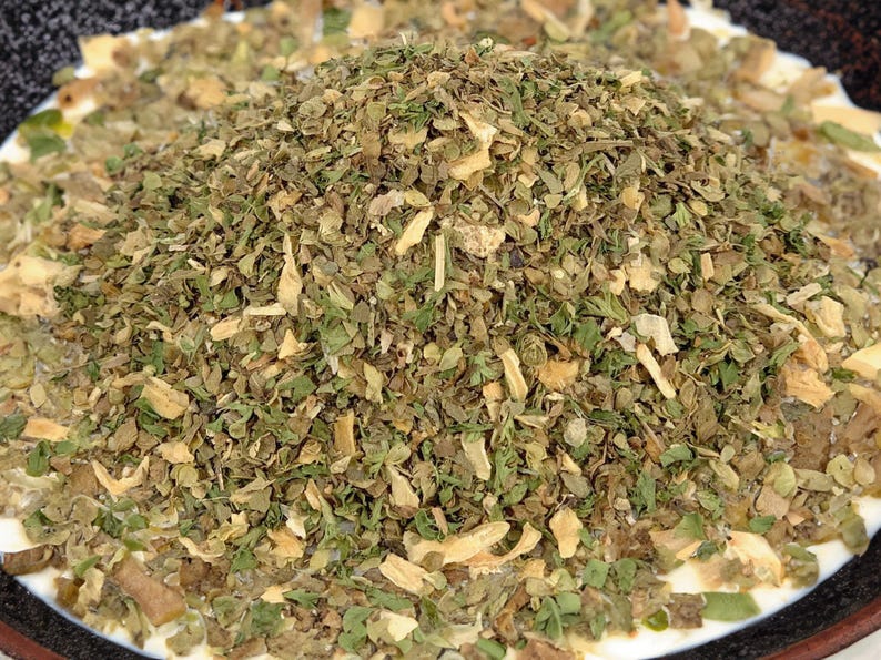 May include: A mound of dried herb seasoning consisting of small, fragmented green leaves and light tan, flaky pieces. The mixture features a variety of textures and shades of olive green, dark green, and pale beige, creating a coarse, dry blend.