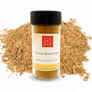 May include: A clear glass jar of Elote Seasoning with a black lid, and a label that reads "Old Town Spice Shop". The seasoning is a mix of brown and green spices. The text on the label says "Sprinkle over roasted corn and enjoy!"