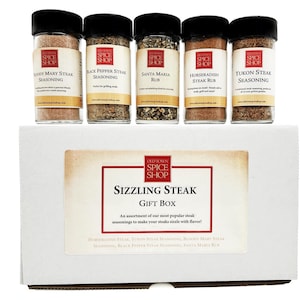 May include: A white gift box with a red and white label that says "Old Town Spice Shop" and "Sizzling Steak Gift Box". The box contains five small glass jars of steak seasoning with labels that say "Bloody Mary Steak Seasoning", "Black Pepper Steak Seasoning", "Santa Maria Rub", "Horseradish Steak Rub", and "Yukon Steak Seasoning".