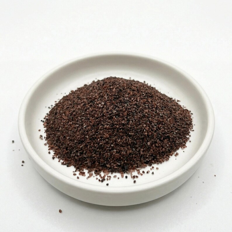 May include: A white ceramic bowl, shallow and round, containing a pile of dark brown, granular substance. The material resembles coarse salt or a spice, with a reddish tinge. The bowl is placed against a plain white backdrop.