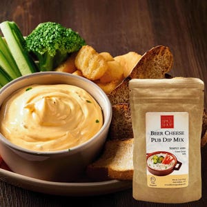 May include: A brown paper bag with a white label that reads "The Original Spice Shop Beer Cheese Pub Dip Mix" with a picture of a bowl of dip and vegetables. The bag is sitting on a wooden table with a bowl of cheese dip, bread, and vegetables.
