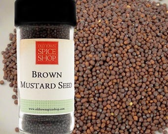 May include: A clear plastic spice jar with a black lid containing brown mustard seeds. The label features a red square with the text Old Town Spice Shop and the words Brown Mustard Seed. A pile of loose brown mustard seeds sits nearby.