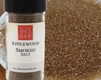 May include: A clear plastic spice jar with a black lid containing brown Applewood Smoked Salt. The label features the text Old Town Spice Shop, Applewood Smoked Salt, and the website address. A pile of the coarse brown salt sits beside the jar.