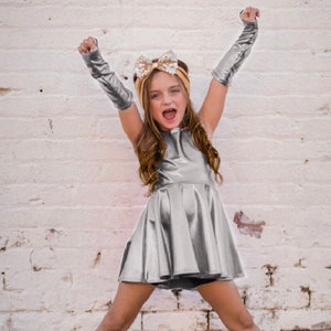 May include: A young girl wearing a silver sequined dress and pink glitter boots poses with her arms raised. She is wearing a gold sequined headband with a bow.