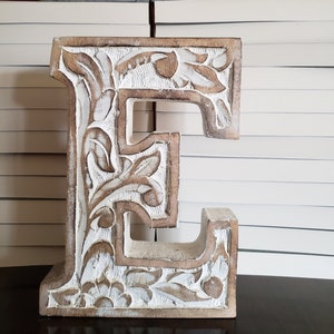 May include: White wooden letter E with intricate floral carvings. The letter is sitting on a stack of white books.