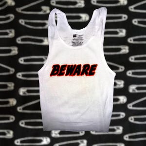 Puede incluir: Camiseta sin mangas blanca con la palabra "BEWARE" en letras rojas y negras. La camiseta sin mangas está hecha de un material acanalado. El fondo es negro con un patrón de imperdibles blancos.