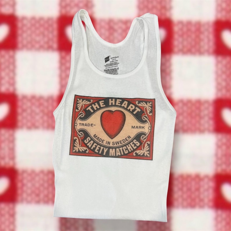 Pode incluir: Camisola branca de al&ccedil;as com um gr&aacute;fico vintage de uma caixa de f&oacute;sforos com o texto "The Heart Trade-Mark Made in Sweden Safety Matches" e um cora&ccedil;&atilde;o vermelho.