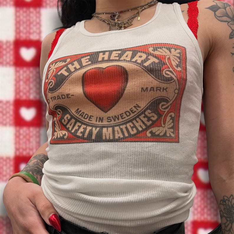 Pode incluir: Camisola branca de al&ccedil;as com um gr&aacute;fico de cora&ccedil;&atilde;o vermelho e o texto "THE HEART MARK TRADE- MADE IN SWEDEN SAFETY MATCHES".