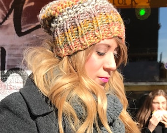 May include: A multi-colored knit beanie in shades of orange, yellow, and gray. The hat is ribbed and has a folded brim. The person is wearing a gray coat and scarf.