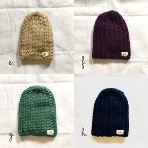 May include: Four crocheted beanies in different colors: beige, purple, green, and navy blue. Each beanie has a small, white label with a symbol on it.