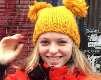 May include: A woman wearing a yellow knit beanie with two pom-poms on top. She is wearing a red jacket and has blonde hair.