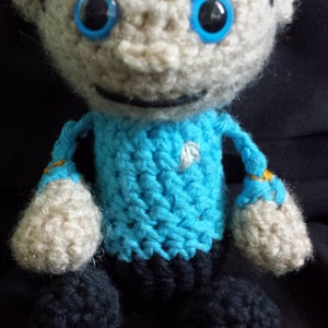 May include: A crocheted toy figure of a person wearing a blue shirt with a white star. The figure has a white face with black eyes and a black line for a mouth.