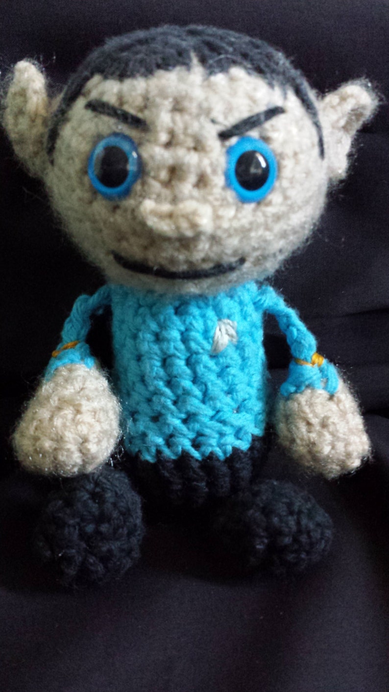 May include: A crocheted toy figure of a person wearing a blue shirt with a white star. The figure has a white face with black eyes and a black line for a mouth.