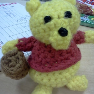 May include: A crocheted Winnie the Pooh bear toy with a red sweater and a brown crocheted bag. The bear is yellow and has black eyes and a black nose.