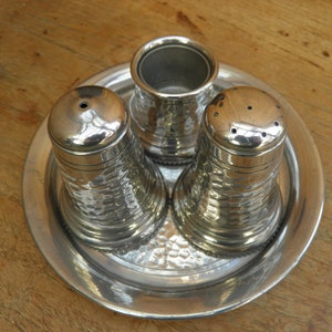 May include: A silver salt and pepper set with a matching container for spices, all resting on a round tray. The set has a hammered texture, and the salt and pepper shakers have multiple holes on top.