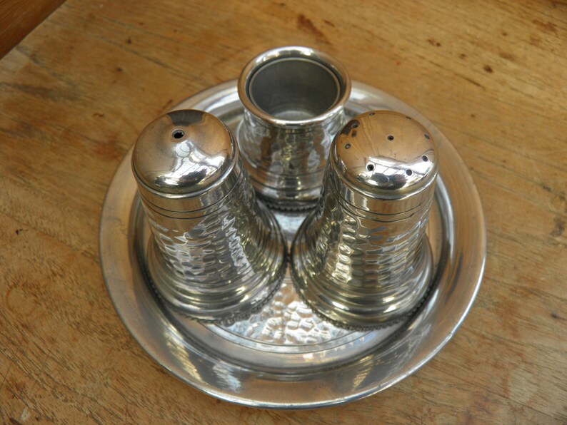 May include: A silver salt and pepper set with a matching container for spices, all resting on a round tray. The set has a hammered texture, and the salt and pepper shakers have multiple holes on top.