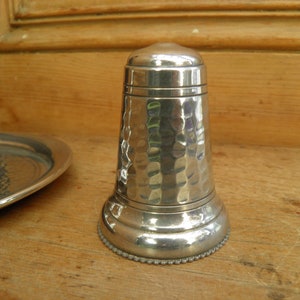 May include: A silver-colored, hammered metal salt shaker with a rounded top and base. The base has a beaded edge. A matching piece and a small tray are visible in the background. The objects are on a wooden surface.
