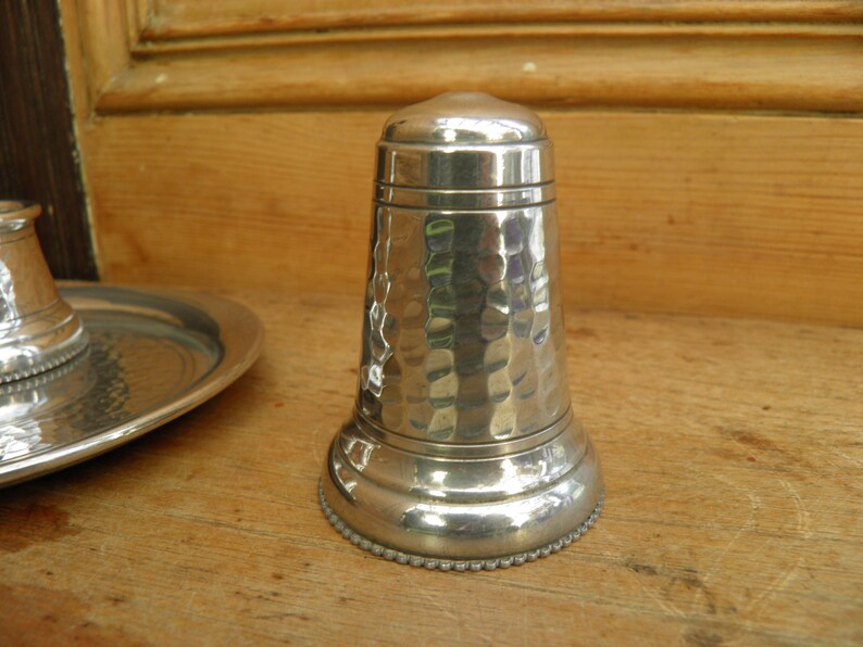 May include: A silver-colored, hammered metal salt shaker with a rounded top and base. The base has a beaded edge. A matching piece and a small tray are visible in the background. The objects are on a wooden surface.