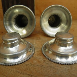 May include: Four antique silver-toned inkwells and pen holders. The inkwells have a round base with a decorative beaded edge and a small, round lid. The pen holders are cylindrical with a flared top.