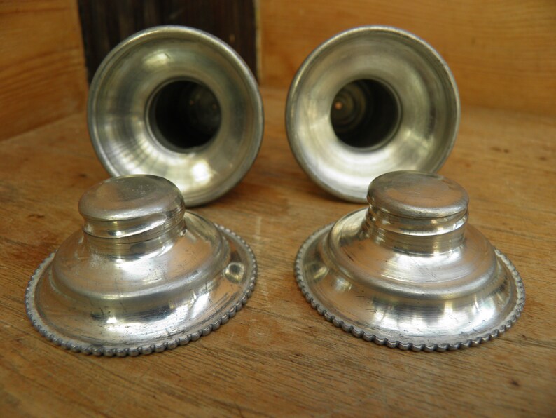 May include: Four antique silver-toned inkwells and pen holders. The inkwells have a round base with a decorative beaded edge and a small, round lid. The pen holders are cylindrical with a flared top.