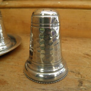 May include: A pair of silver salt and pepper shakers with a hammered texture. The salt shaker has a series of small holes on top. The base of the shakers have a beaded design. The shakers are on a wooden surface.