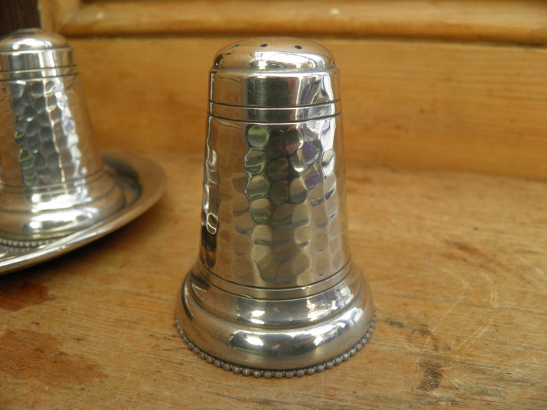 May include: A pair of silver salt and pepper shakers with a hammered texture. The salt shaker has a series of small holes on top. The base of the shakers have a beaded design. The shakers are on a wooden surface.