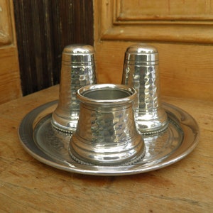 May include: A silver salt and pepper shaker set with a hammered texture, and a matching open container, all resting on a round tray. The set is displayed on a wooden surface, suggesting a rustic or vintage aesthetic.