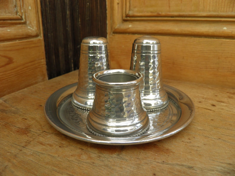 May include: A silver salt and pepper shaker set with a hammered texture, and a matching open container, all resting on a round tray. The set is displayed on a wooden surface, suggesting a rustic or vintage aesthetic.