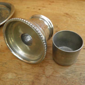 May include: A collection of silver metal serving pieces, including a small cup and a decorative pedestal with a hammered texture. The pedestal has a beaded edge. The pieces are on a wooden surface.