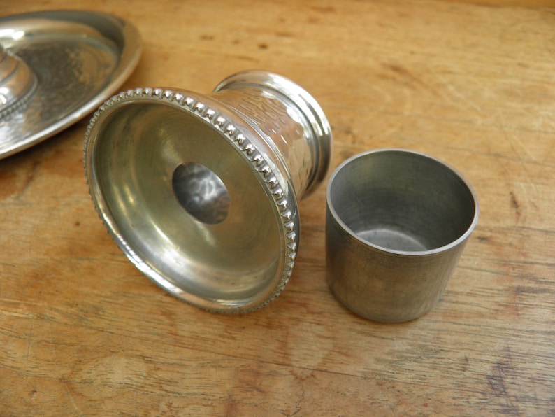 May include: A collection of silver metal serving pieces, including a small cup and a decorative pedestal with a hammered texture. The pedestal has a beaded edge. The pieces are on a wooden surface.