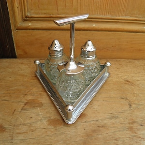 Puede incluir: Un juego de sal y pimienta vintage plateado con una bandeja triangular. El juego incluye dos saleros de vidrio con tapas plateadas y un recipiente central de vidrio. La bandeja tiene un borde con cuentas y un asa.