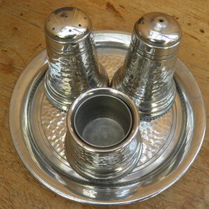 May include: A silver salt and pepper shaker set with a matching container, presented on a round tray. The set has a hammered texture, and the tray has a decorative border. The items are made of metal and sit on a wooden surface.