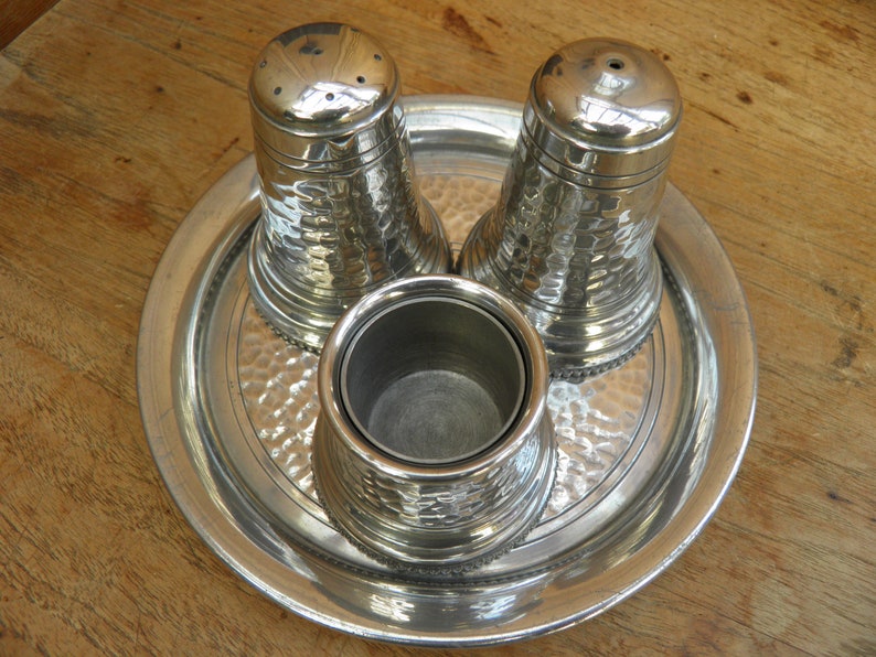 May include: A silver salt and pepper shaker set with a matching container, presented on a round tray. The set has a hammered texture, and the tray has a decorative border. The items are made of metal and sit on a wooden surface.