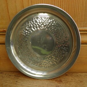 May include: A round, silver-colored metal tray with a hammered texture in the center and a smooth, raised rim. The tray has a circular design with a central, smooth area. The overall design is simple and elegant.