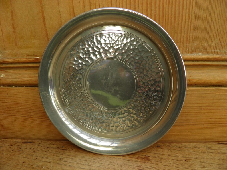 May include: A round, silver-colored metal tray with a hammered texture in the center and a smooth, raised rim. The tray has a circular design with a central, smooth area. The overall design is simple and elegant.