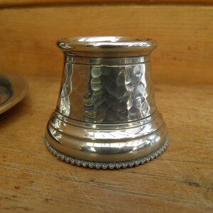 May include: A silver-colored, hammered metal candle holder with a textured surface. The base has a beaded edge. The candle holder is cylindrical with a flared base and a rounded top. The background is a wooden surface.