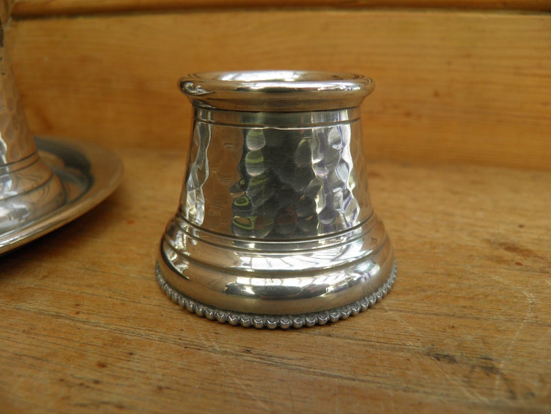 May include: A silver-colored, hammered metal candle holder with a textured surface. The base has a beaded edge. The candle holder is cylindrical with a flared base and a rounded top. The background is a wooden surface.