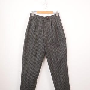 May include: High-waisted trousers in a dark gray and black micro-check pattern. These pants feature a structured waistband with a single button closure, front pleats, and a tapered leg silhouette with cuffed hems, hanging on a wooden hanger.