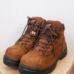 May include: Brown leather work boots with black soles and brown laces. The boots have a high-top design and are on a light wooden surface. The Red Wing logo is visible. The boots are suitable for construction or outdoor work.