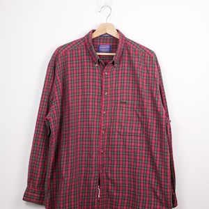 May include: A red and green plaid button-down shirt hanging on a wooden hanger. The shirt has long sleeves and a button-down collar. The Pendleton label is visible at the top. The shirt is made of a soft, woven fabric.