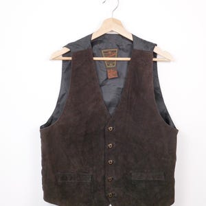 May include: A brown suede vest with a V-neck and button closure. The vest has two front pockets and is hanging on a wooden hanger. The interior lining is dark gray.