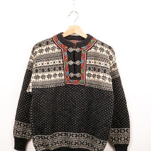 Peut inclure: Un pull à manches longues gris anthracite avec un empiècement crème orné de motifs de flocons de neige noirs. L'encolure présente une fermeture à agrafes en métal argenté entourée d'une bordure brodée de fleurs colorées.