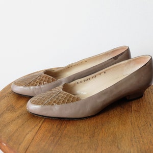 May include: A pair of taupe low-heeled pumps featuring a textured, reptilian-patterned leather toe cap. The shoes have a smooth, solid-colored heel and side panel, a rounded toe, and a low, block-style heel. The interior lining is a light beige.
